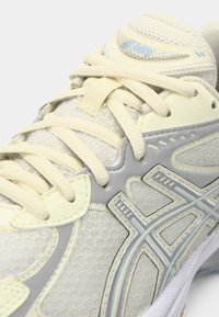 Ανοιχτό γκρι αθλητικό παπούτσι με πλέγμα και συνθετικά υλικά. Διαθέτει εκρού κορδόνια, υφασμένο πάτο και μπλε λεπτομέρειες στον πλαϊνό σχεδιασμό.