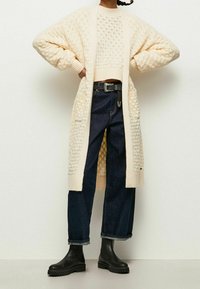 Krémový pletený kardigan ke kolenům s otevřeným designem, spárovaný s tmavě modrými džínami s vysokým pasem a černými kotníkovými botami.