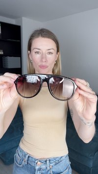 Zwarte oversized zonnebril met een gradient bruin glas, glanzende afwerking en patroon op de pootjes, vastgehouden voor een neutrale achtergrond.
