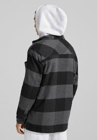 Man som vänder ryggen i en svart och grå rutad jacka med en ljusgrå huvtröja under mot en enkel grå bakgrund.