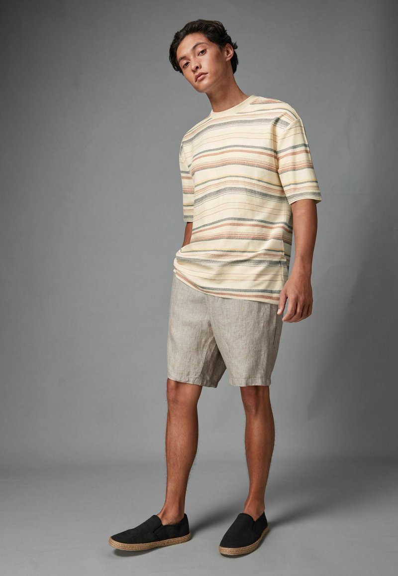 Randig kortärmad skjorta i kräm, beige och pastellfärger, matchad med ljusgrå shorts och svarta slip-on skor med en strukturerad yta.