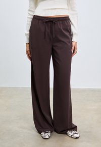 Weite Hosen aus dunkelbraunem Stoff mit einer glatten Textur, ausgestattet mit einem elastischen Bund und einer Kordel. Kombiniert mit gemusterten Schuhen.