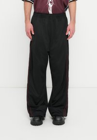 Μαύρο αθλητικό παντελόνι από δίχτυ με ελαστική μέση, διακοσμημένο με κόκκινες πλαϊνές ρίγες και άνετη εφαρμογή. Λεπτομέρειες από υφή αεριζόμενου υφάσματος.