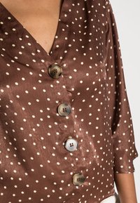 Brun blus med prickar och V-halsringning samt tre stora, glänsande knappar. Tillverkad av ett mjukt tyg med en något glänsande yta. Korta ärmar.