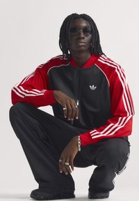 Čierna a červená športová bunda na zips s bielymi pruhmi a logom, skombinovaná s čiernymi nohavicami a tmavou obuvou. Doplnky zahŕňajú prstene a náramok.