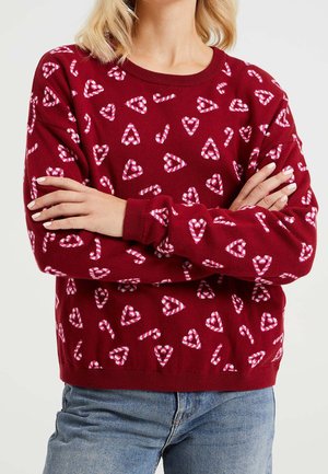 Femme portant un pull rouge avec des cœurs en sucre d'orge roses et blancs, bras croisés, associée à un jean en denim bleu.