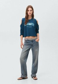 Teal skrajšan majica s prozračnimi rameni, bela grafična besedila, v kombinaciji s širokimi svetlimi kavbojkami in vzorčastim pasom. Ravne sandale.