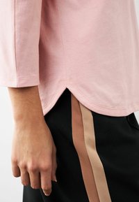 Top de algodón rosa claro de manga larga con dobladillo curvado y aberturas laterales, combinado con pantalones negros con rayas marrones.