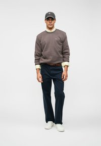 Man in een grijze sweatshirt, beige shirt, marineblauwe broek, witte sneakers en een donkergrijze pet staat tegen een effen witte achtergrond.