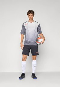 Noor noormängija halli ja valge särgiga, mustade lühikeste pükstega, valgete sokkidega ja spordijalatsitega hoiab jalgpalli ühetaolise tausta ees.