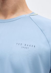 Κοντινό πλάνο ανοιχτό μπλε αθλητικής μπλούζας με το λογότυπο "TED BAKER SPORT" στο στήθος, που φοριέται από ένα άτομο.