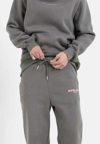 Sudadera gris y pantalones de chándal a juego; tela suave. La sudadera tiene un cuello redondo y un logo rosa, y los pantalones tienen un cordón ajustable.