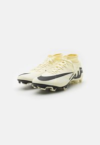 Botas de fútbol de color amarillo claro con una superficie texturizada, acentos negros y un diseño de cuello alto; presentan una suela con tacos para mayor tracción.