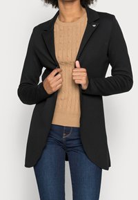 Czarna, dopasowana marynarka o gładkiej strukturze, z otwartym przodem i subtelnymi klapami, noszona na jasnobrązowym swetrze w warkocze. Uzupełniona o małą przypinkę.