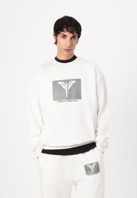 Hvid sweatshirt med et grafisk print og sorte diagonale striber samt teksten "CARLO COLUCCI". Kombineret med matchende hvide bukser.