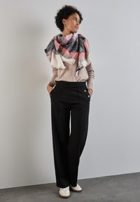 Pantaloni neri con due bottoni decorativi, abbinati a un maglione marrone chiaro e a una sciarpa grande a quadri multicolori. Tessuto liscio, vestibilità casual.