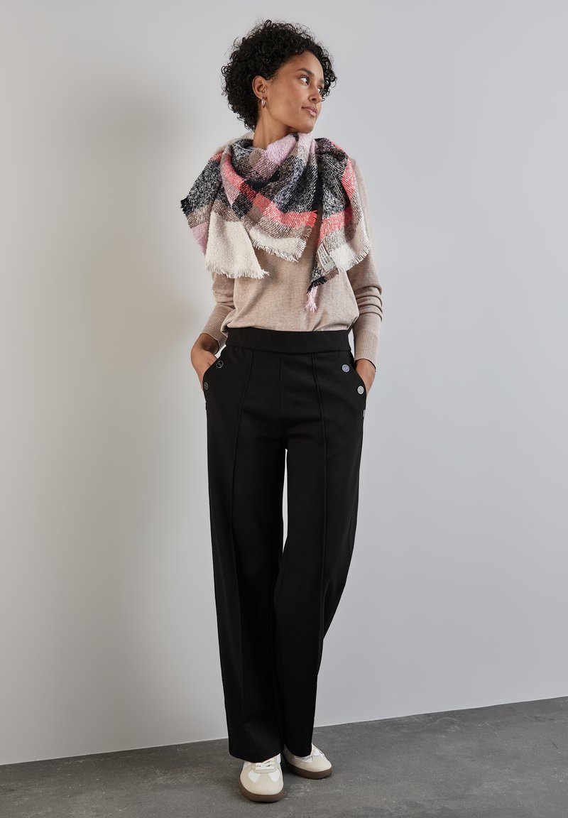 Pantaloni neri con due bottoni decorativi, abbinati a un maglione marrone chiaro e a una sciarpa grande a quadri multicolori. Tessuto liscio, vestibilità casual.