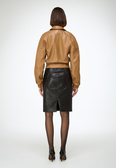 Barna bőr bomber dzseki túlméretezett ujjakkal, fekete bőr ceruzaszoknyával, amely hátsó sliccel rendelkezik, átlátszó fekete harisnyával viselve.