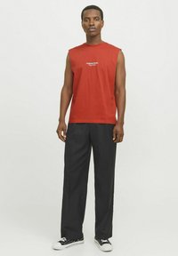Rød ærmeløs t-shirt med et logo på brystet, parret med sorte vidde bukser og sorte sneakers. Glat stof og afslappet design.