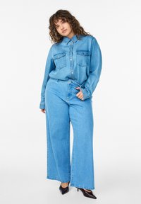 Žena s kučeravými vlasmi v svetlomodrej rifľovej košeli a džínsach so širokými nohavicami pózuje, pričom rukou sťahuje slučku na opasku, na nohách má čierne špicaté lodičky.