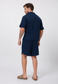 Marineblaues Kurzarmhemd und passende Shorts aus strukturiertem Stoff. Verfügt über einen klassischen Kragen und seitliche Schlitze. Sandalen runden den Look ab.