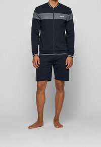 Marinblå träningsoverall med grå rand över bröstet, dragkedjestängning, ribbad muddar och matchande shorts. Har en vit logotyp på bröstet.