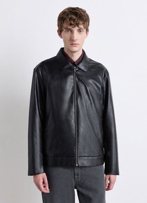 Junger Mann mit kurzem lockigem Haar, der eine schwarze Lederjacke mit Reißverschluss und dunkelgraue Hose vor einem schlichten hellen Hintergrund trägt.