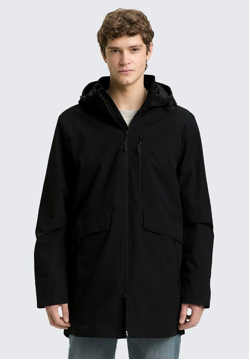 Noor mees musta lukuga kapuutsiga jope ja taskutega, seisab heledal hallil taustal.