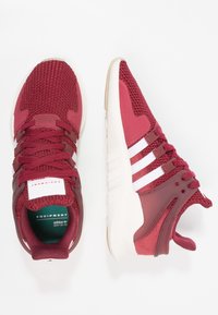 Bordové sportovní boty s texturovaným pleteným svrškem, bílými pruhy, polstrovanou podrážkou a zpevněnou patou. Lehký a prodyšný design.