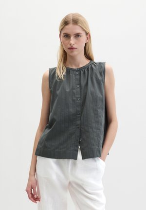 Młoda kobieta o blond włosach, ubrana w ciemnoszarą, bezrękawną bluzkę na guziki i białe spodnie, stoi z jedną ręką w kieszeni.