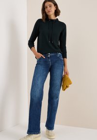 Dunkelgrüner Kapuzenpullover mit Kordeln, blaue weiter geschnittene Jeans, beigen Sneakern und einer gefalteten senfgelben Strickmütze.