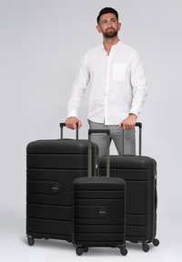 Hombre con camisa blanca sosteniendo las asas de tres maletas negras de diferentes tamaños sobre un fondo gris claro liso.