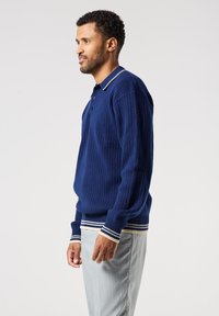 Námořnický pulovr s ribovaným vzorem a polo límcem, vybavený pruhovaným lemem a manžetami v bílé a béžové barvě. Látka se zdá být texturovaná a měkká.