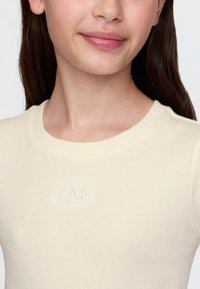 Jaunas asmuo su ilgais tamsiais plaukais, dėvintis kremu ribinetą marškinių su užsiuvimu „GAP“ ant krūtinės, rodo švelnią šypsnį.