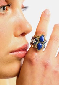 Bague en or avec deux pierres en forme de goutte d'eau bleues, ornée de perles colorées et de motifs complexes, portée sur un doigt près d'un visage.