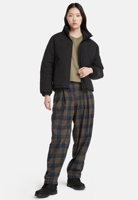 Giubbotto piumino nero con colletto alto, indossato sopra una camicia oliva. Pantaloni a quadri larghi in brown e blu, abbinati a stivali neri.