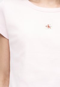 Κοντινό ενός ατόμου που φοράει ροζ ριμπ κοντομάνικη μπλούζα με μικρό κόκκινο λογότυπο Calvin Klein στο στήθος.