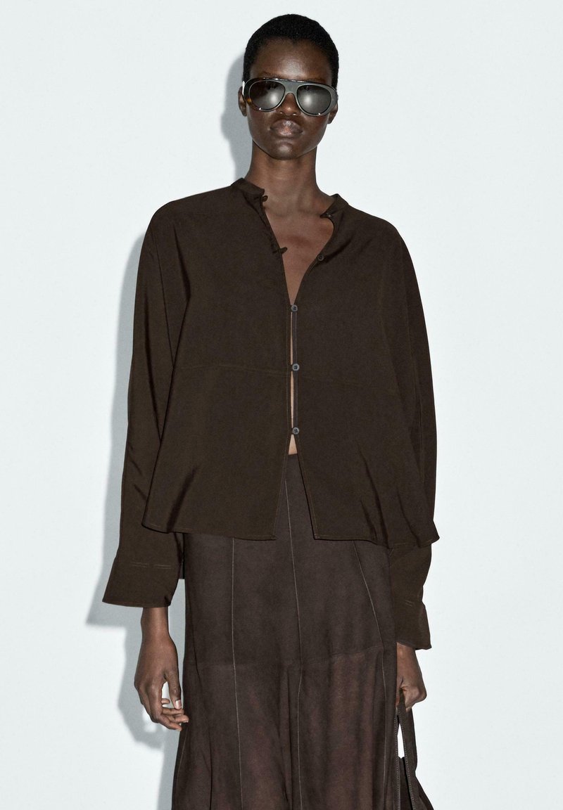 Brun stof knap-uppet bluse med en rund krave og løs pasform, parret med en matchende nederdel. Model iført oversized solbriller.