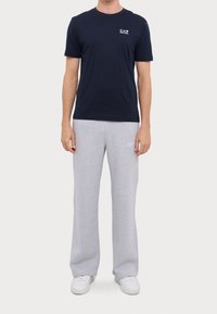 Man i en marinblå EA7 t-shirt, ljusgrå träningsoverall med liten vit text, och vita sneakers, som står mot en enkel bakgrund.