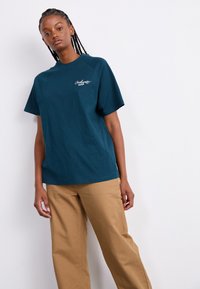 Teal cotton T-shirt with short raglan sleeves and a crew neckline, featuring a white embroidered logo on the left chest. Paired with beige pants.