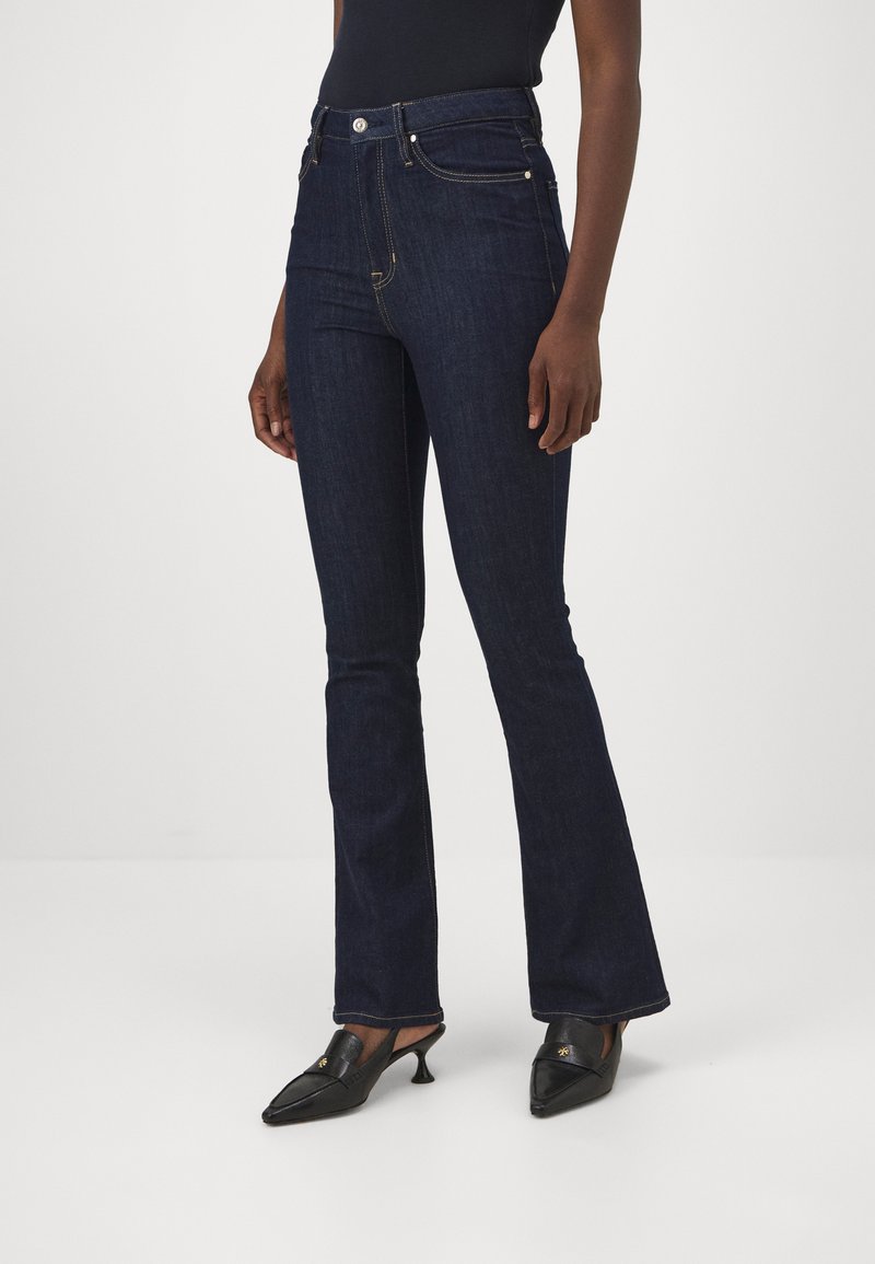 Jeans acampanados de denim oscuro con cintura alta, costuras visibles y detalles de botones de metal. Combinados con zapatos negros de punta afilada.