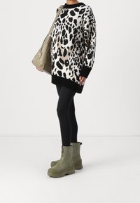 Person klädd i en överdimensionerad tröja med svart och vitt djurtryck, svarta leggings, olivgröna stövlar och bärande en olivgrön axelväska.