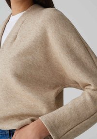 Maglione lavorato a maglia beige con un fit rilassato, caratterizzato da un collo a V, maniche lunghe e una texture morbida. Il tessuto presenta un delicato motivo melangiato.
