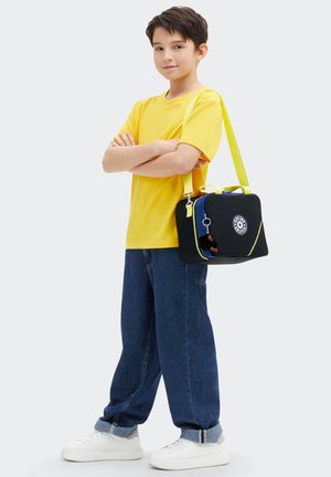 Sac noir avec des accents jaune fluo, fermeture éclair bleue et patch rond. Porté à l'épaule par une personne en chemise jaune et en jean bleu.