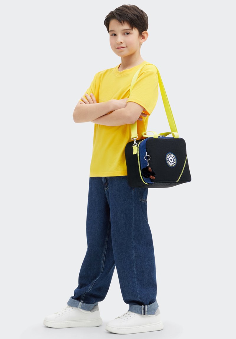 Sac noir avec des accents jaune fluo, fermeture éclair bleue et patch rond. Porté à l'épaule par une personne en chemise jaune et en jean bleu.