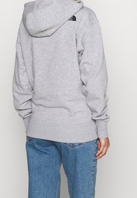 Šedá mikina s kapucí, ribbovanými manžetami a lemem, s malým černým logem na horní části zad, v kombinaci s modrými džínovými kalhotami.