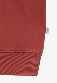 Κόκκινο-καφέ πλεκτό ύφασμα με ριμπ μανσέτα και μια μικρή μπεζ ετικέτα με πορτοκαλί κείμενο στη ραφή πλευράς.