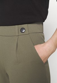 Högerm hand i fickan på hovda olivegröna byxor med svart knapp, kombinerat med en instoppad svart topp.