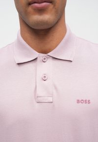 Világos rózsaszín galléros póló texturált anyagból, két gombos patenttal és bal oldali mellrészen hímzett márkajelzéssel.