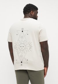 Bež majica iz bombaža z grafiko črnega geometrijskega sonca in lune na hrbtni strani, s trikotnimi elementi in podrobnim risanjem linij.